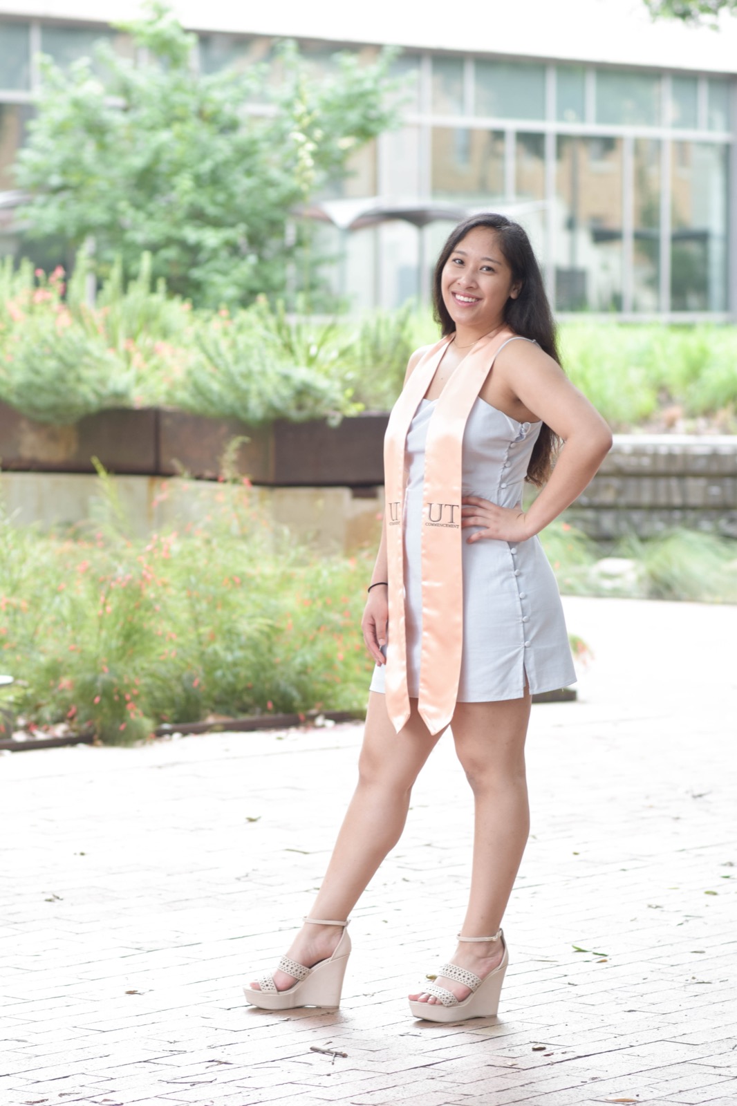 A graduate in a light blue dress and peach commencement stole, hand on hip, smiling on an outdoor walkway.