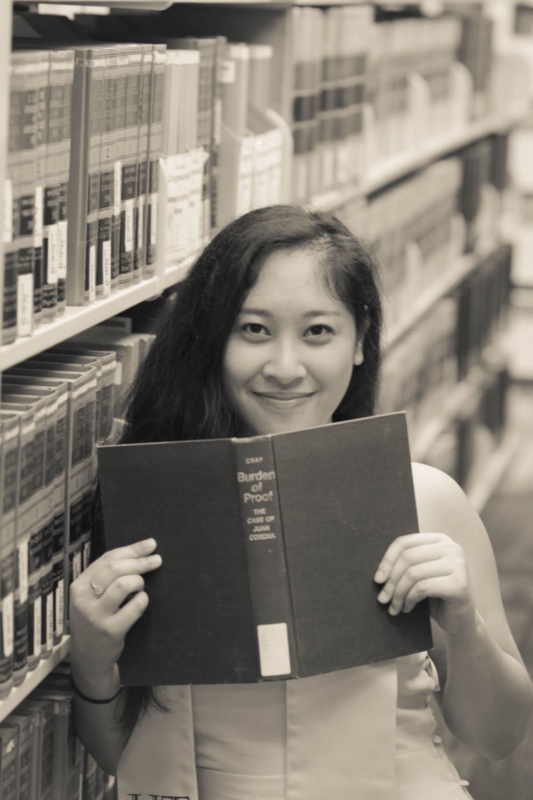 Black-and-white portrait of a woman peeking over an open antique book in library stacks.