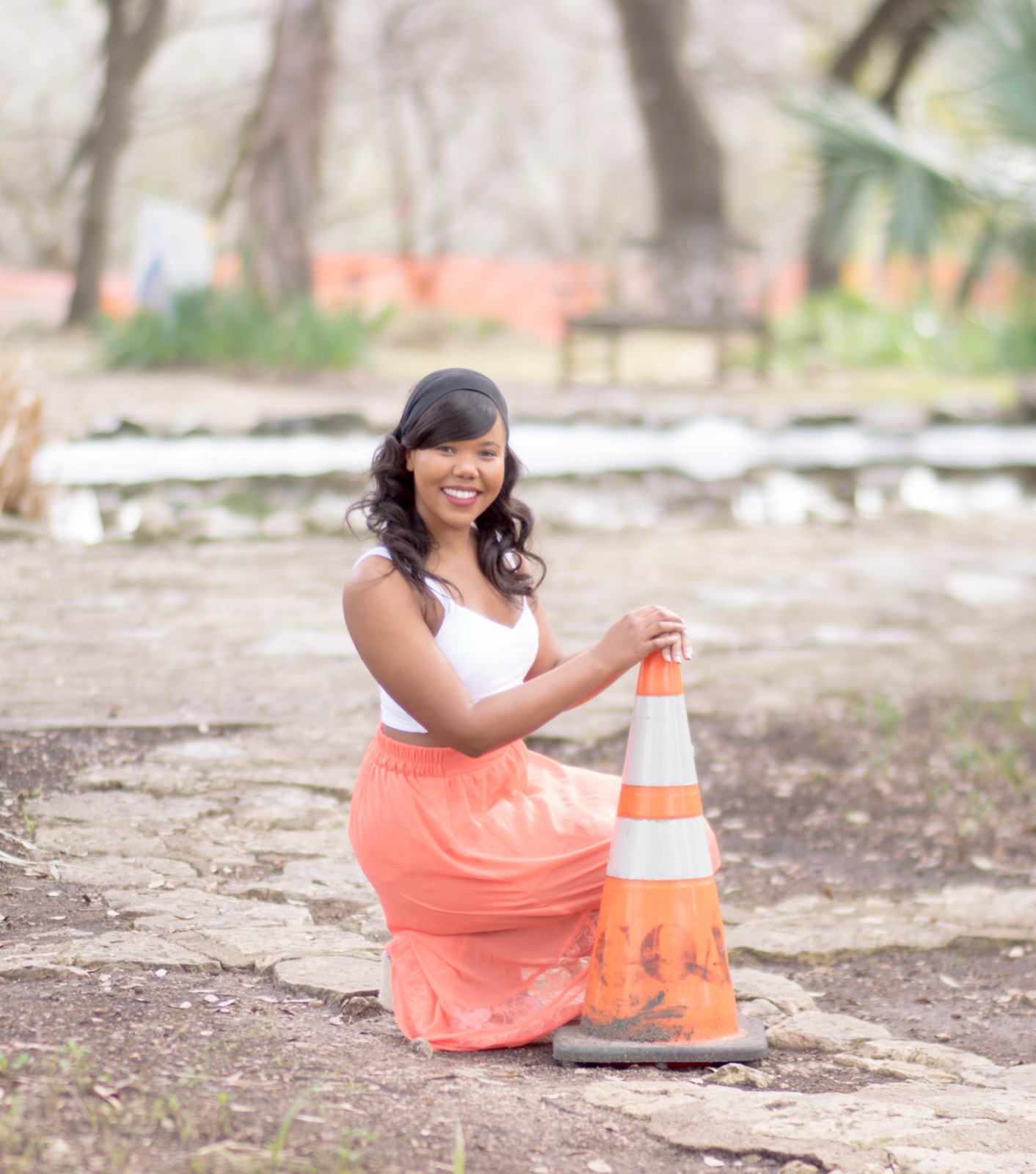 A woman kneels beside a traffic cone on a stone path, smiling, in a coral skirt and white top.