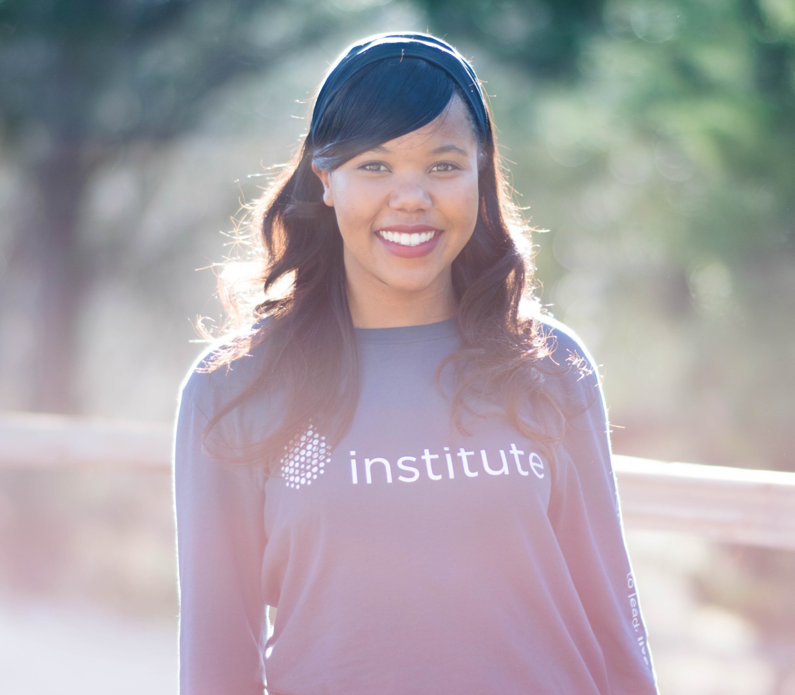 Close, backlit portrait of a smiling woman in a headband and long-sleeve graphic shirt outdoors.